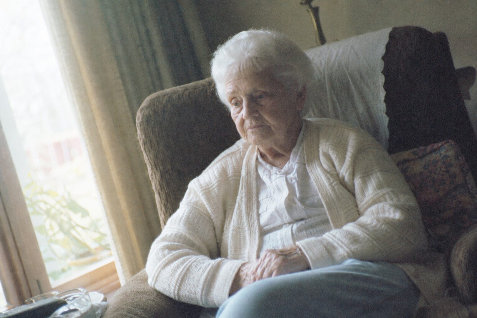 An elderly white-haired woman with her head bowed by a window