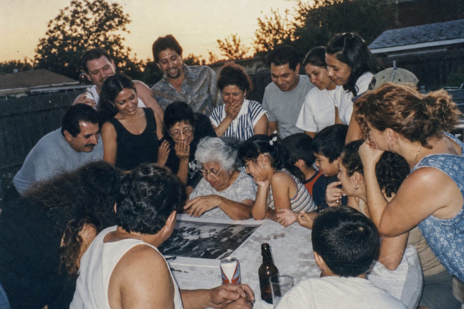 Fourteen family members gathering around a single old photograph in a backyard