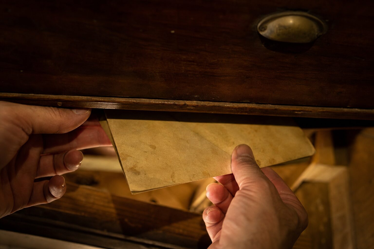 Close-up of hands pulling an old brown envelope from beneath a loose wooden panel at the bottom of a dresser drawer
